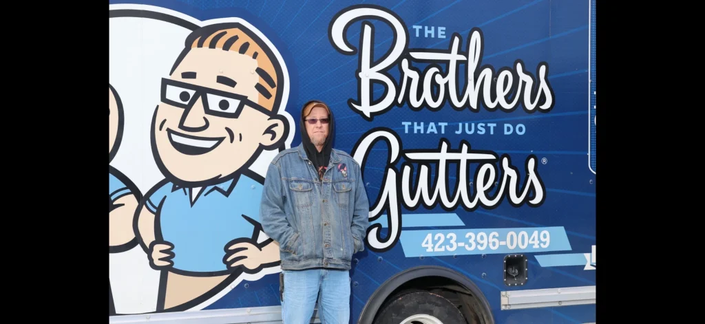 Gutter installation employee standing in front of a blue trailer displaying the company logo and slogan.