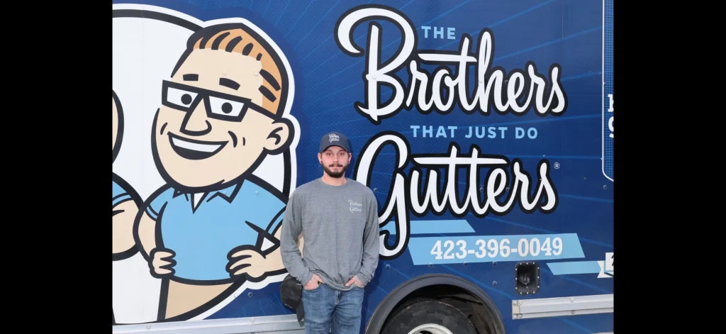 Gutter installation employee standing in front of a blue trailer displaying the company logo and slogan.