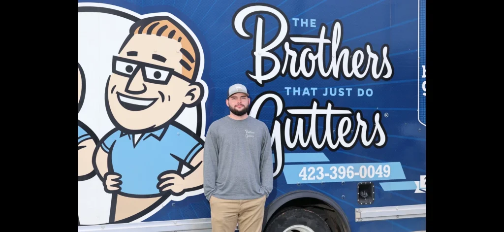 Gutter company team member standing in front of a blue service trailer with a cartoon logo and company slogan.