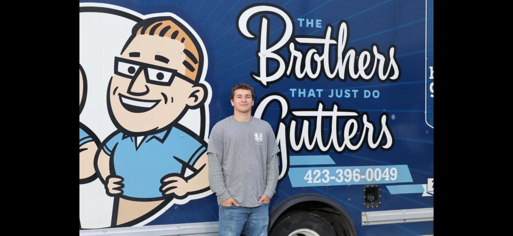 Employee posing in front of a branded gutter service trailer featuring a cartoon character and company phone number.