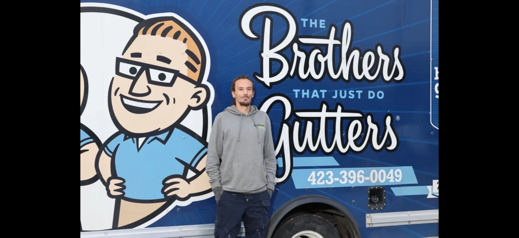 Gutter company team member standing in front of a blue service trailer with a cartoon logo and company slogan.