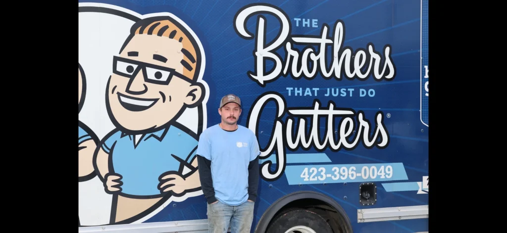 Gutter installation worker standing in front of a blue service trailer with cartoon logo and company slogan.